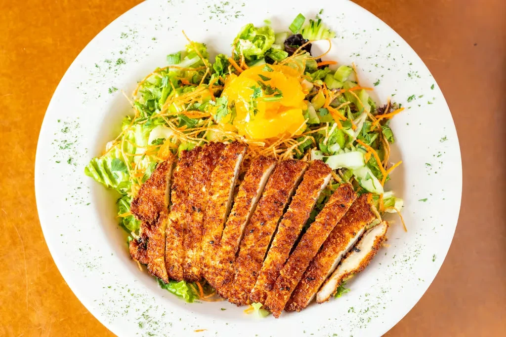 Overhead of sliced fried cutlet, green salad, orange segments on white plate.