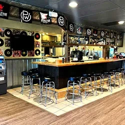 Empty bar interior: long wooden counter, stools, and record-decorated walls.