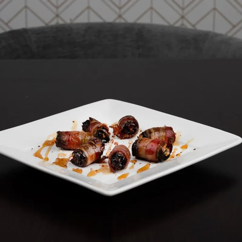 Seven bacon-wrapped dates with glaze on a white square plate.