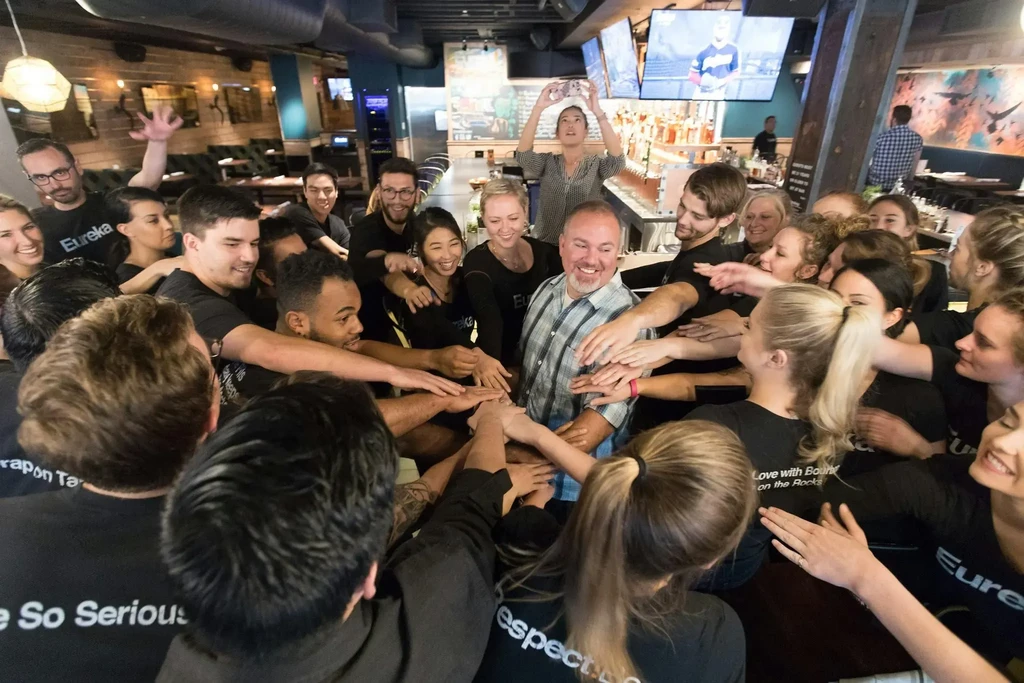 Smiling restaurant staff huddle, hands stacked, inside a bar.