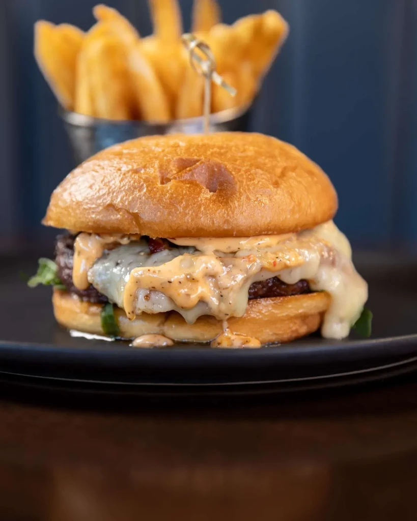 Gourmet burger with brioche bun, melted cheese, orange sauce, and fries.