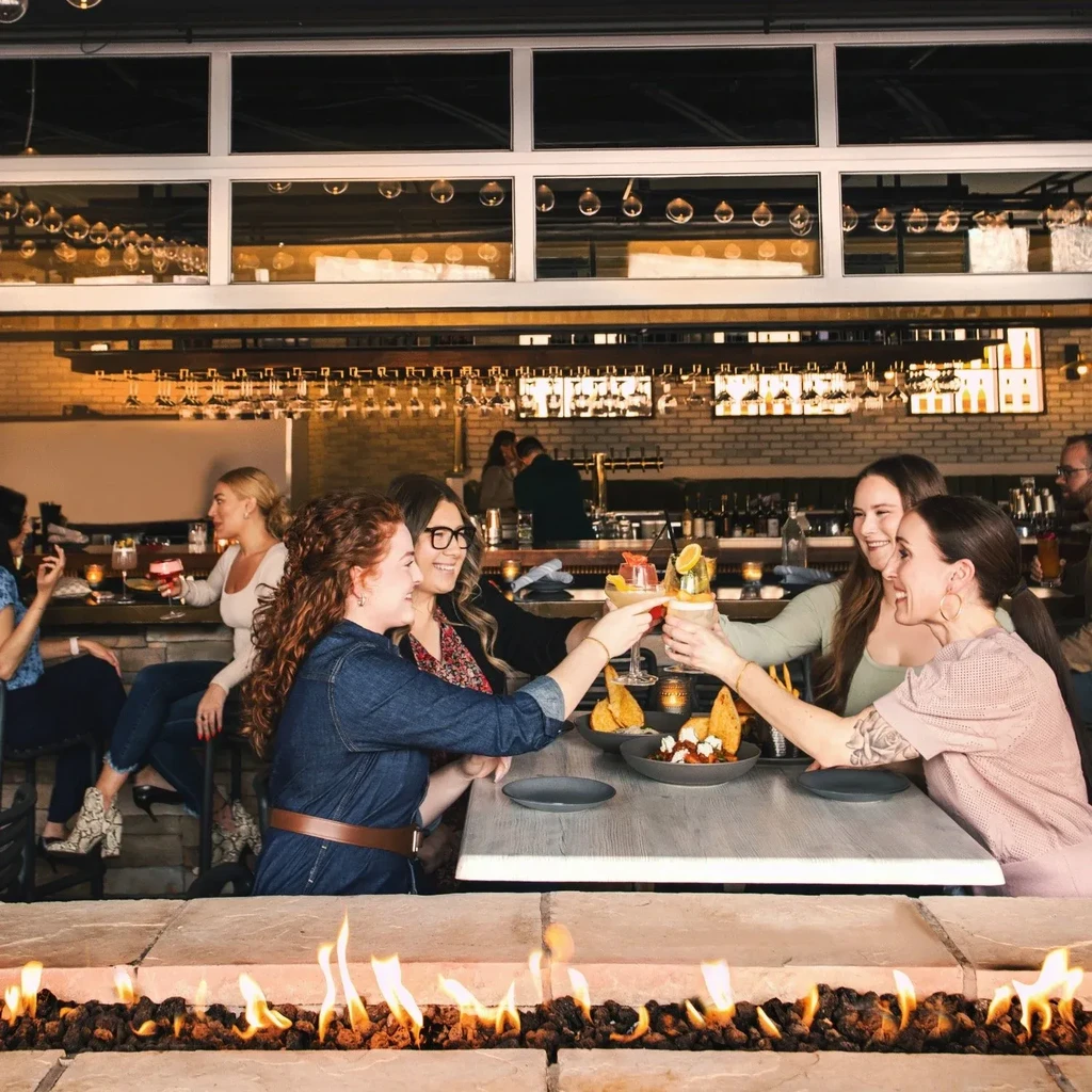 Four women toast colorful cocktails by a restaurant's fire pit.