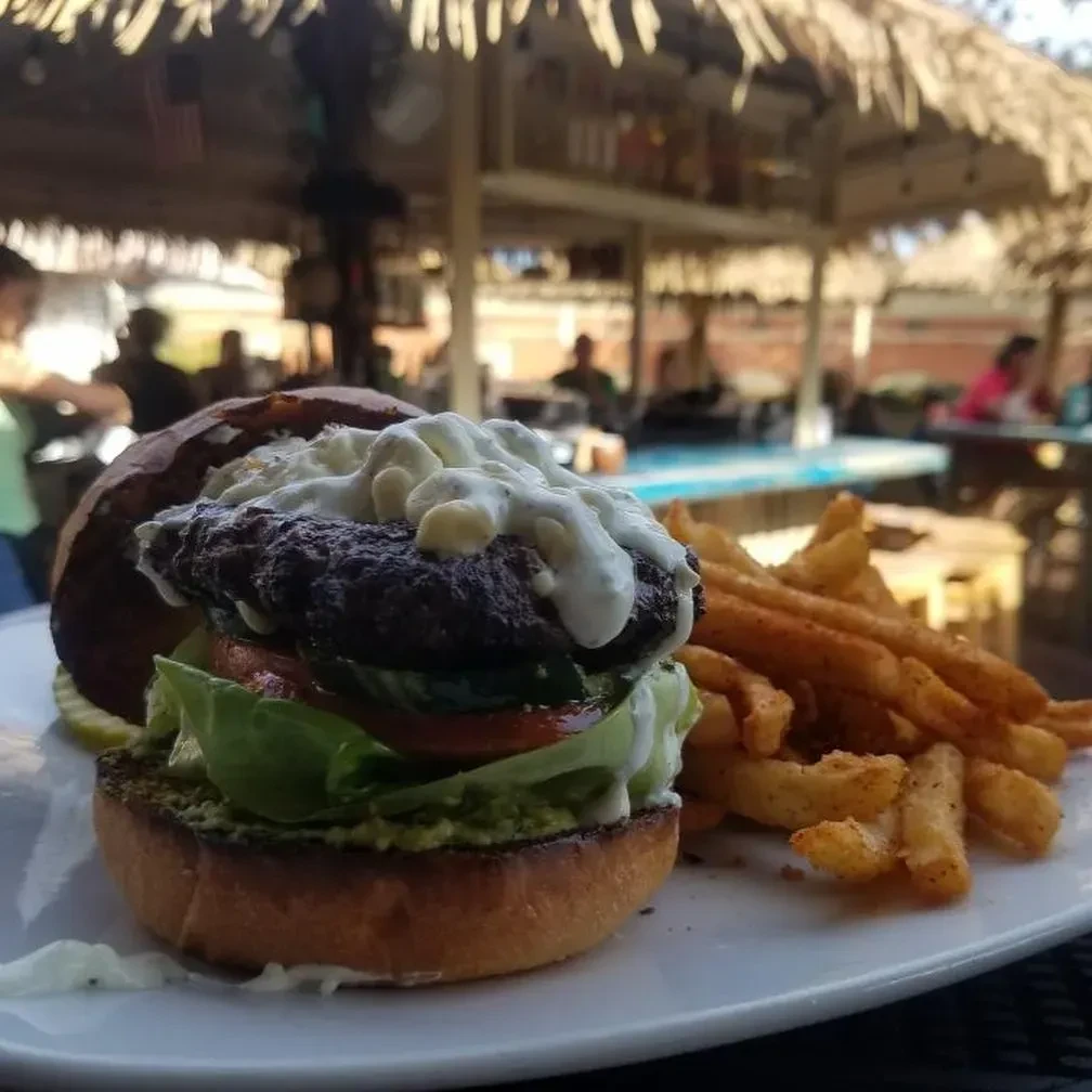 Gourmet burger with macaroni, fries, pickle, and blurred thatched roof.