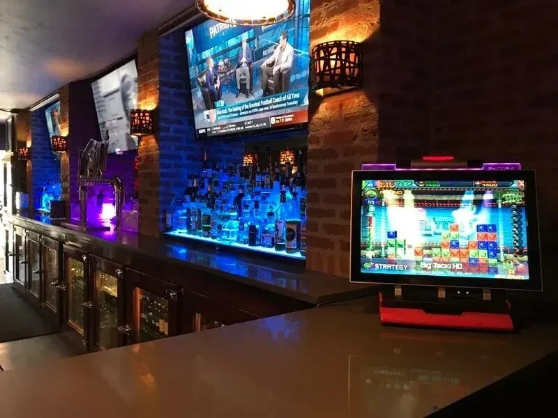 Modern bar interior with blue LED liquor display, TVs, and arcade game.
