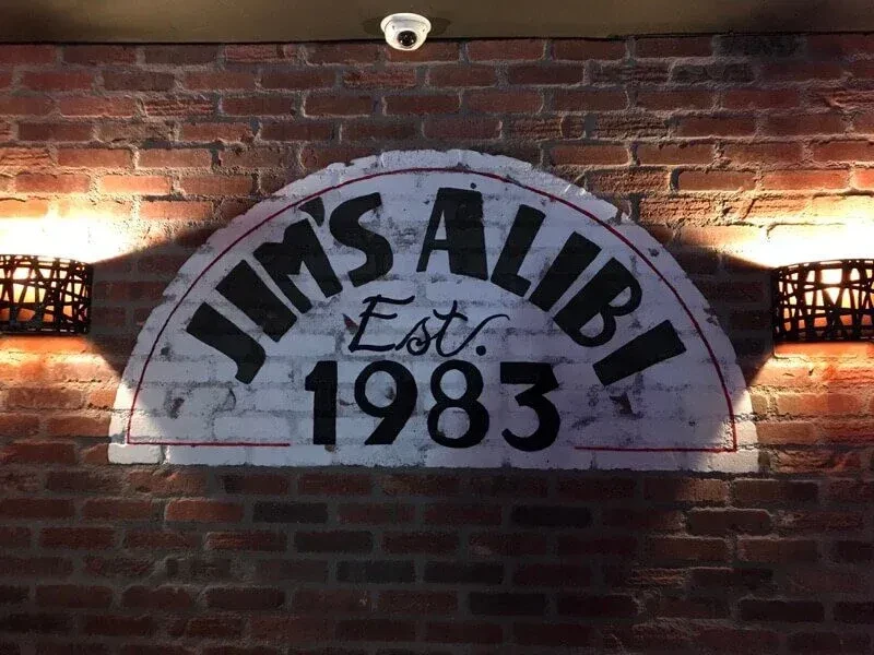 Rustic brick wall with "JIM'S ALIBI Est. 1983" sign and security camera.