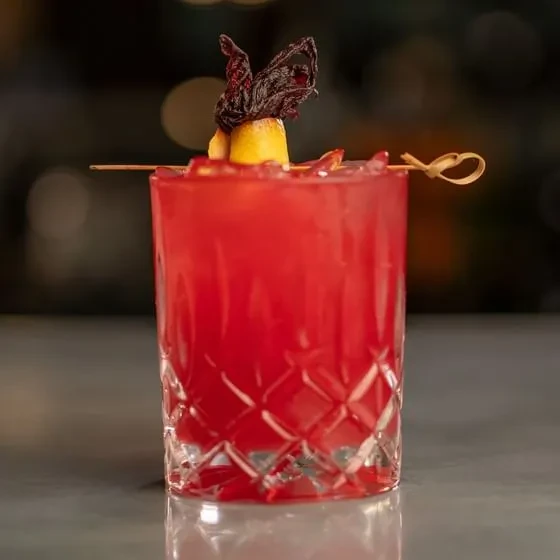 Red cocktail in cut glass with citrus and hibiscus garnish.