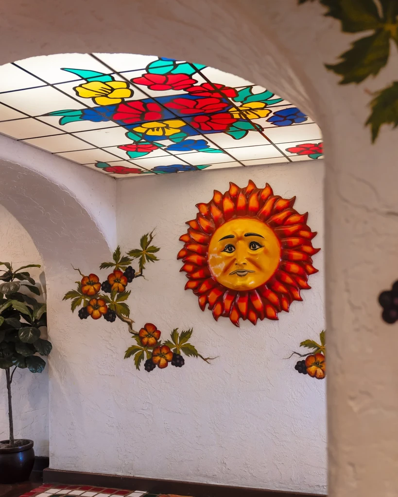 Archway with large 3D sun, wall murals, and colorful stained-glass ceiling.