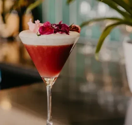 Red cocktail with white foam and pink flowers in a martini glass.