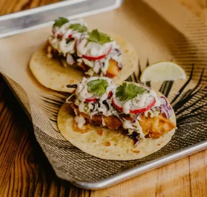 Two golden fish tacos topped with slaw, crema, cilantro, and lime.