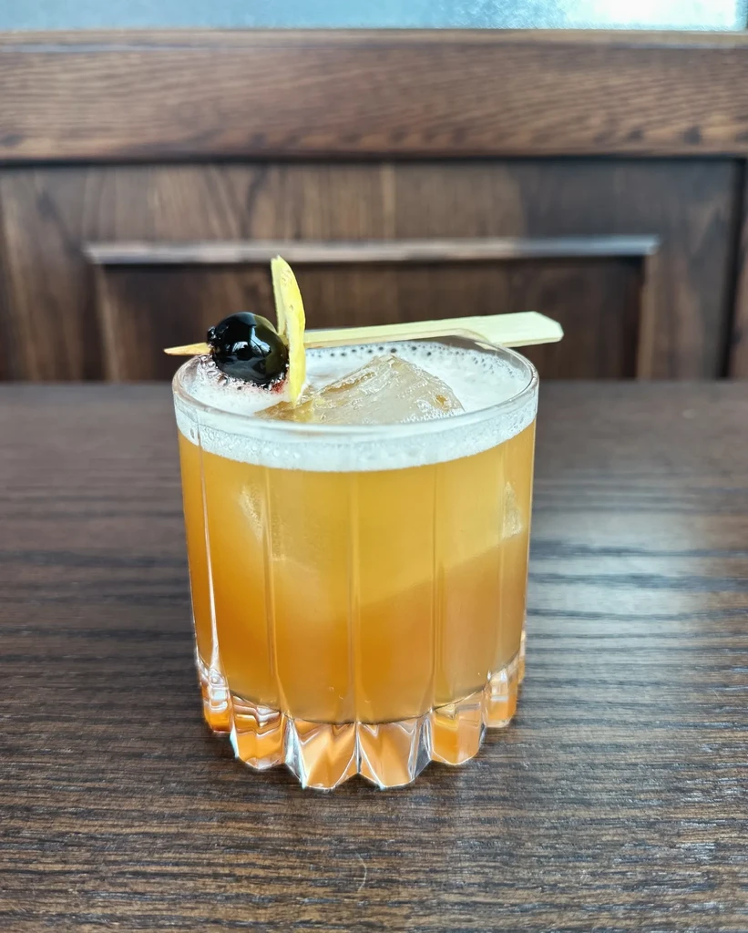 Amber cocktail in faceted glass with foam, cherry, lemon peel, stirrer.