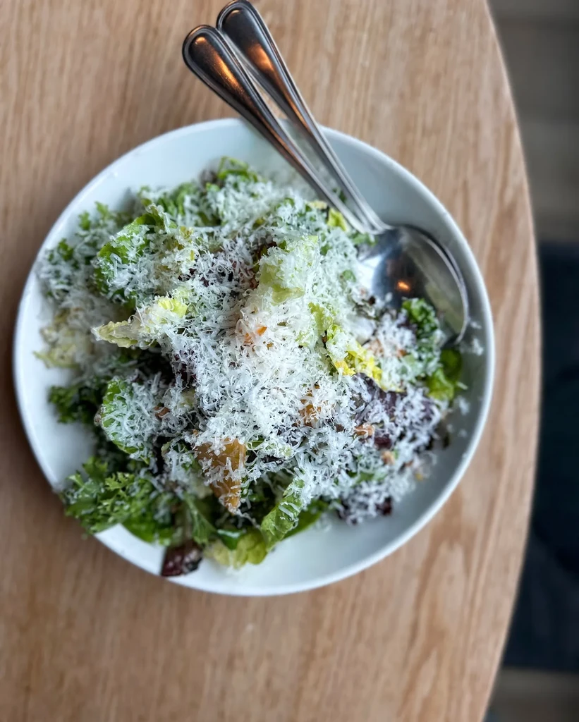 Overhead salad in white bowl with greens, grated cheese, spoons.