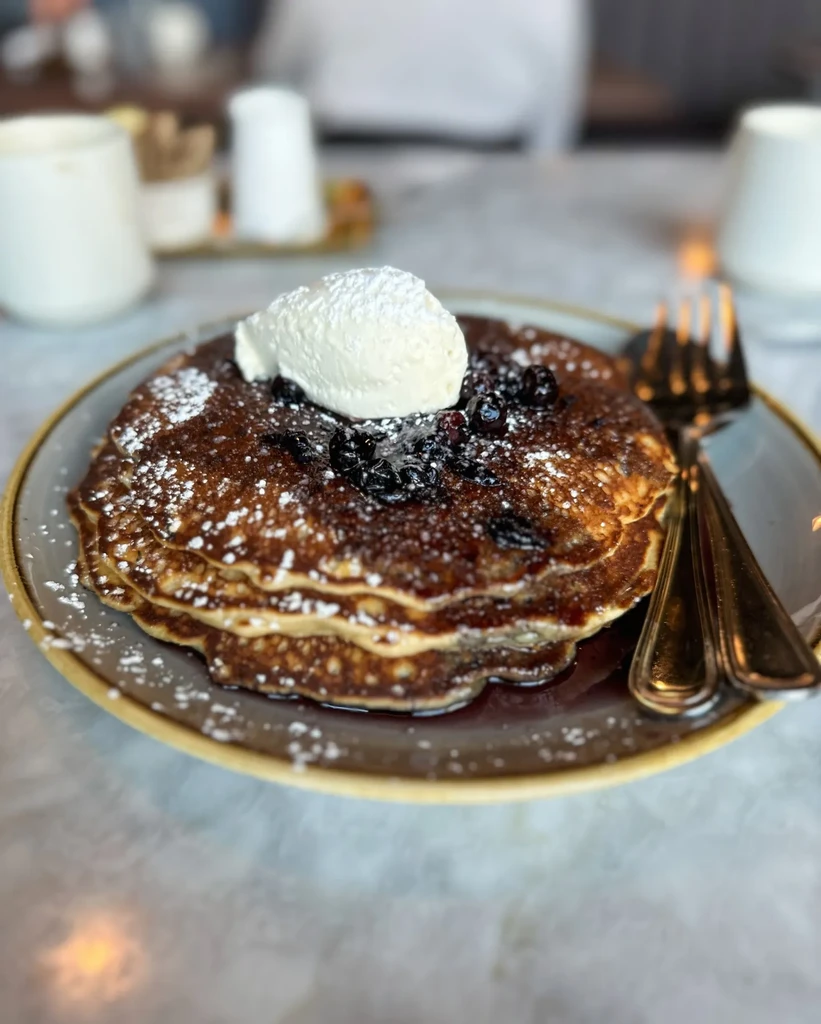 Stack of pancakes with cream, dark berries, powdered sugar, and syrup.