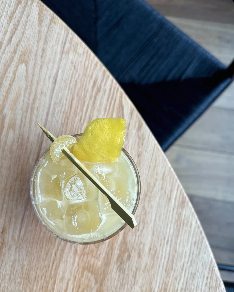 Overhead: Yellow cocktail with lemon and pick on a light wood table.