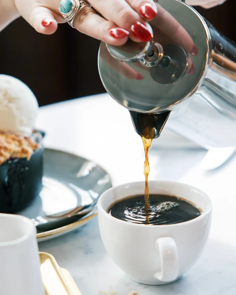 Hand with ring pours coffee from silver French press into white cup.
