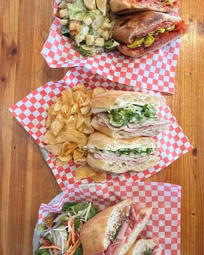 Three deli sandwiches, salads, and chips on a wooden table.