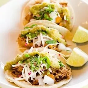 Three shredded meat tacos with vegetables, green and white sauces, lime.