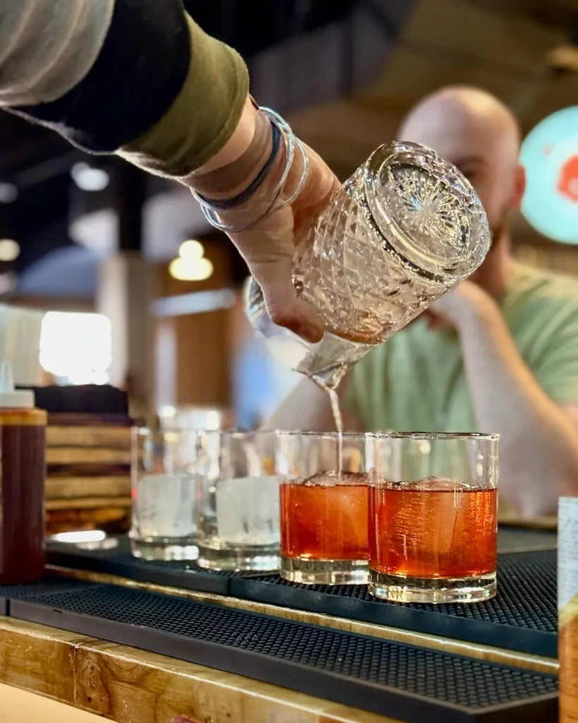 Arm pours clear liquid from pitcher into iced amber drink.