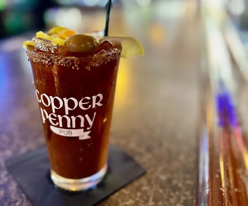Garnished Bloody Mary cocktail in logoed glass on dark bar counter.