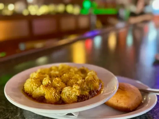 Savory gratin with golden baked topping, a bread roll, and blurred lights.