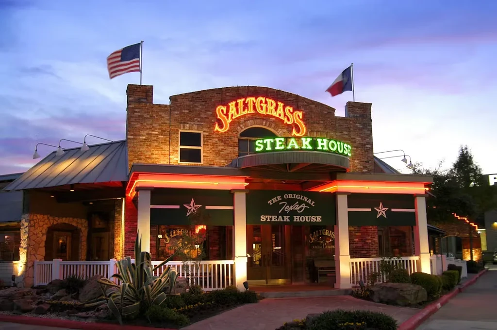 Saltgrass Steak House exterior at twilight, brick building, neon signs, flags.