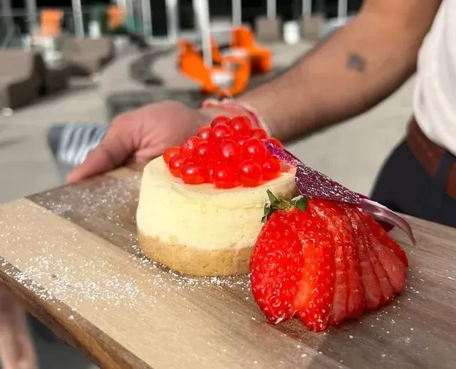 Individual cheesecake with sliced strawberry and red spheres on wooden board.