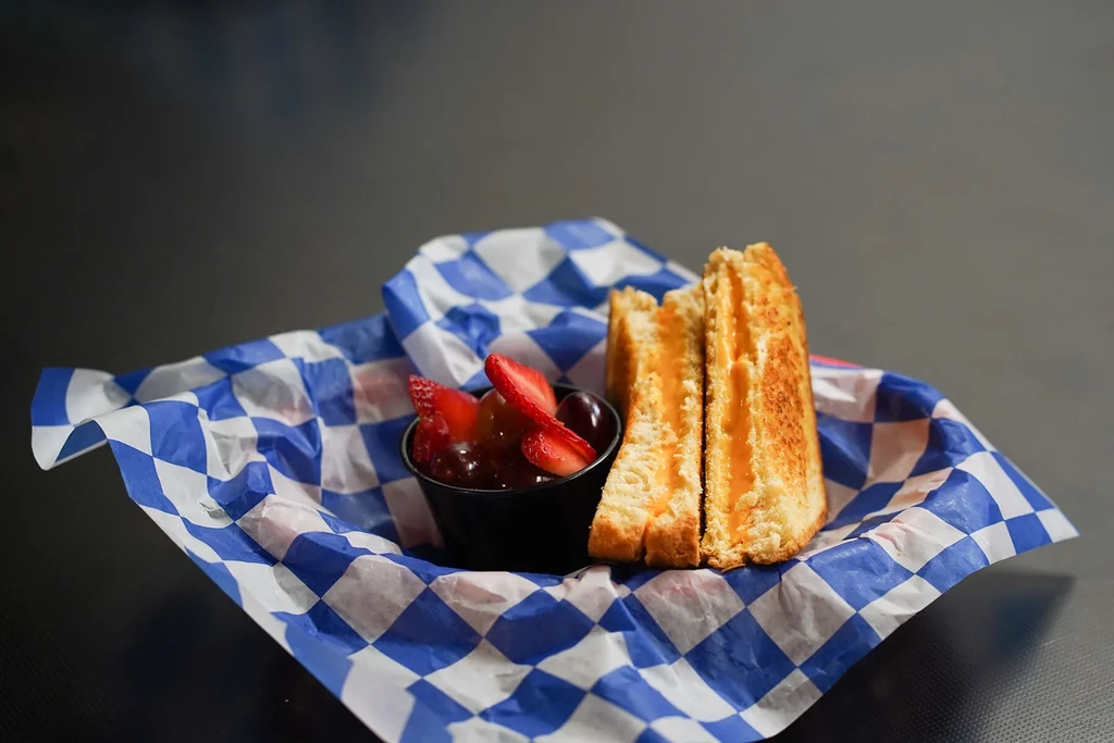 Half grilled sandwich, melted orange filling, fruit cup on checkered paper.