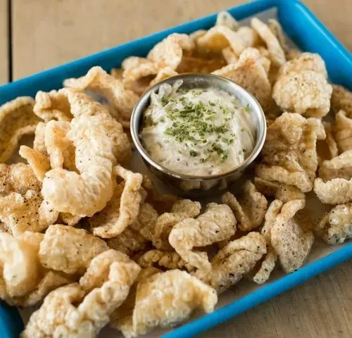 Crispy, seasoned chicharrones with herbed dipping sauce in a blue tray.