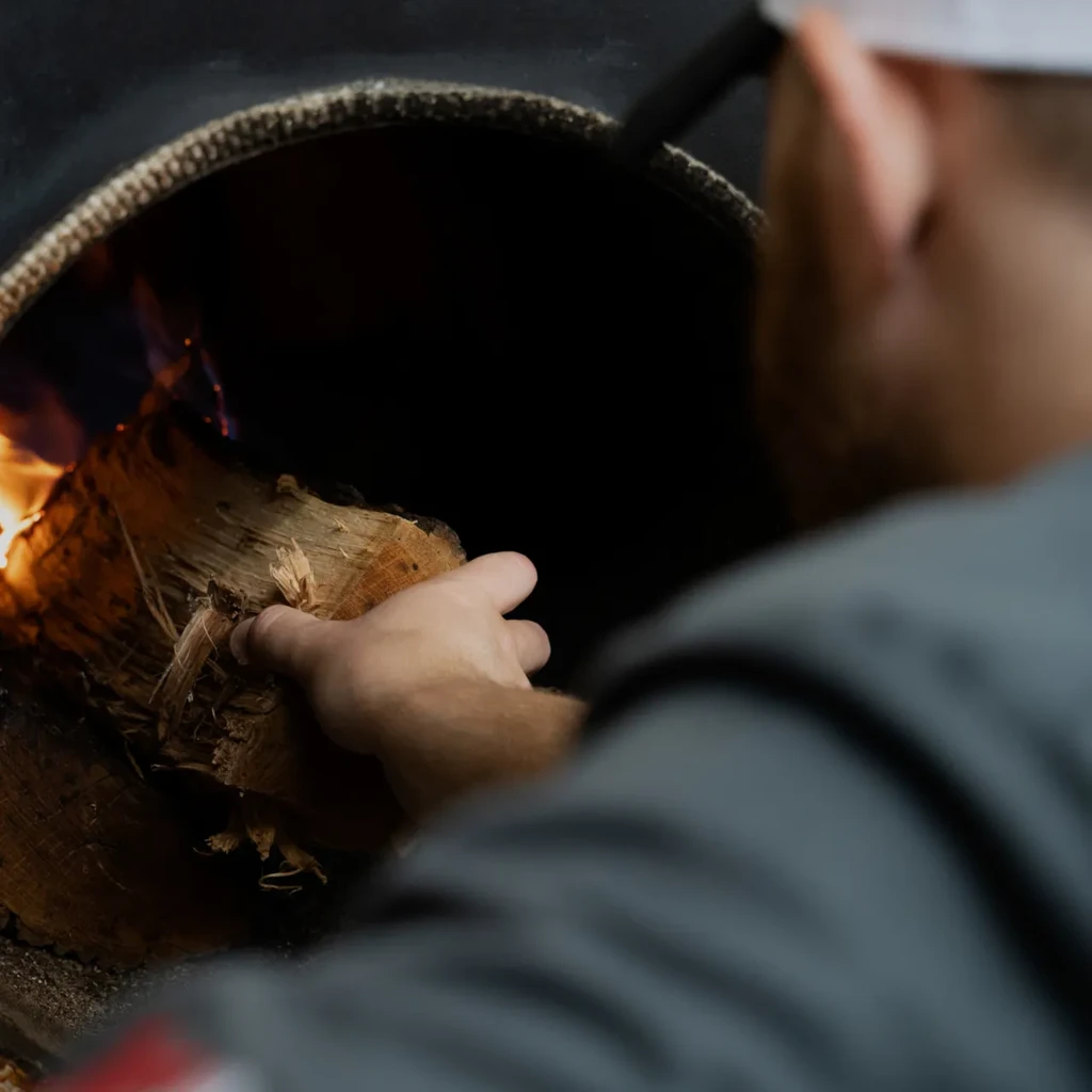 Hand placing firewood into a hot, wood-fired oven with flames.