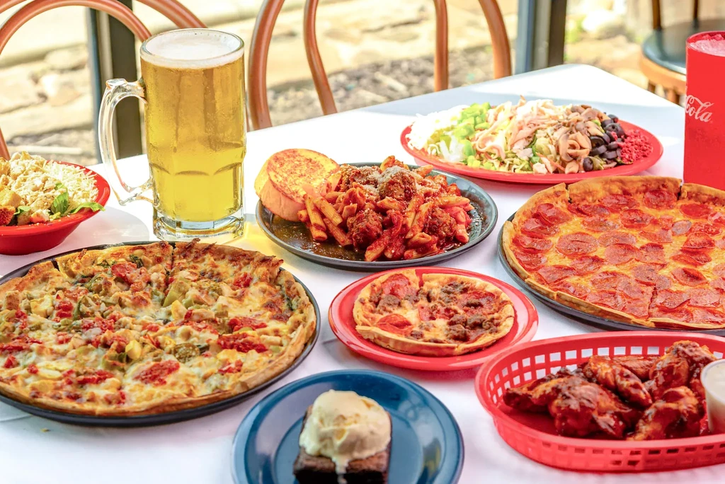 Table spread with pizzas, pasta, wings, salad, and beer.