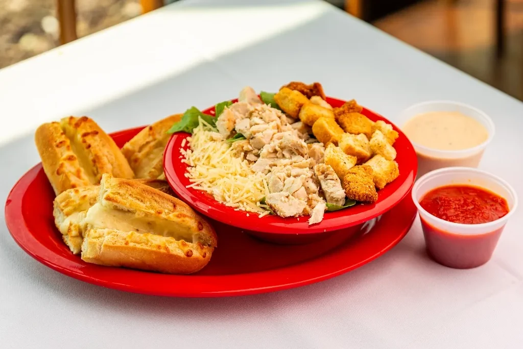 Salad with chicken, breadsticks, and sauces on red plate and bowl.
