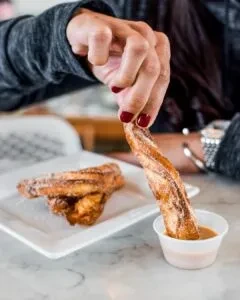 Hand dips a sugar-cinnamon churro into light brown sauce.