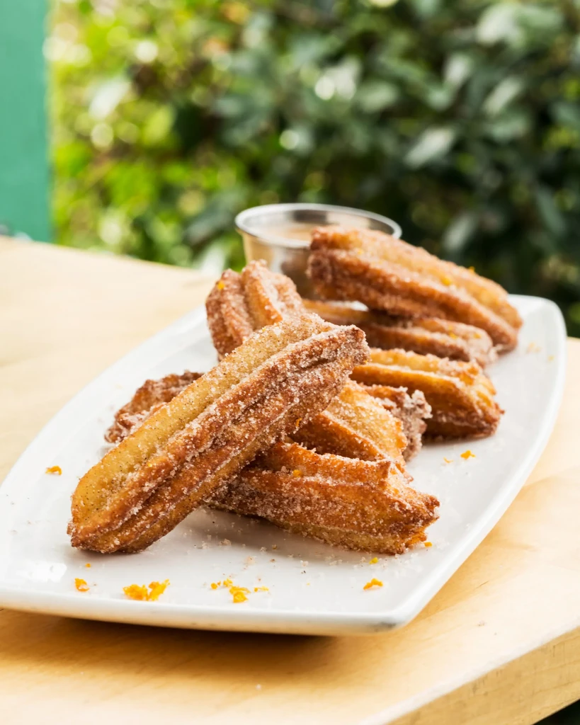 Plate of golden churros with sugar, orange zest, and dipping sauce.