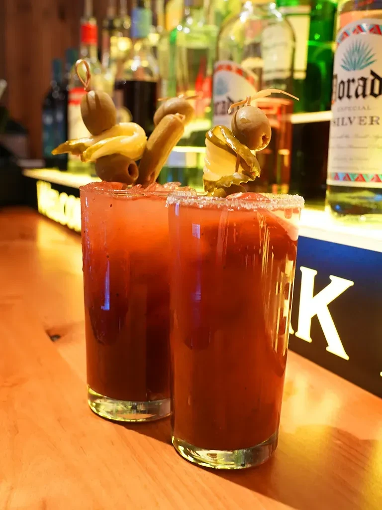 Two garnished Bloody Mary cocktails on a bar with blurred liquor bottles.