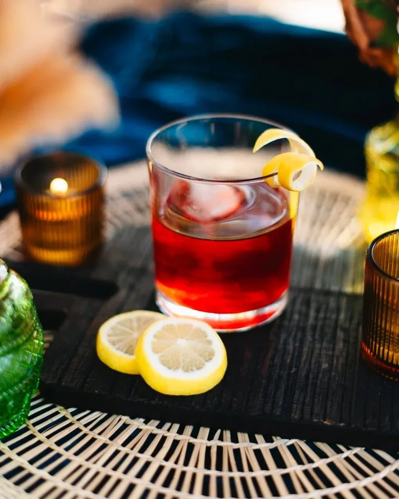 Dark red cocktail with lemon garnish, ice, in a cozy setting.