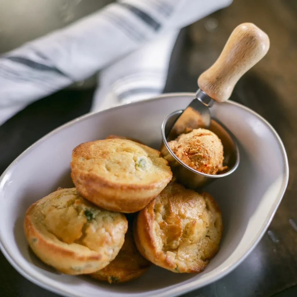 Golden-brown mini-muffins with green bits, red spread, and knife in a bowl.