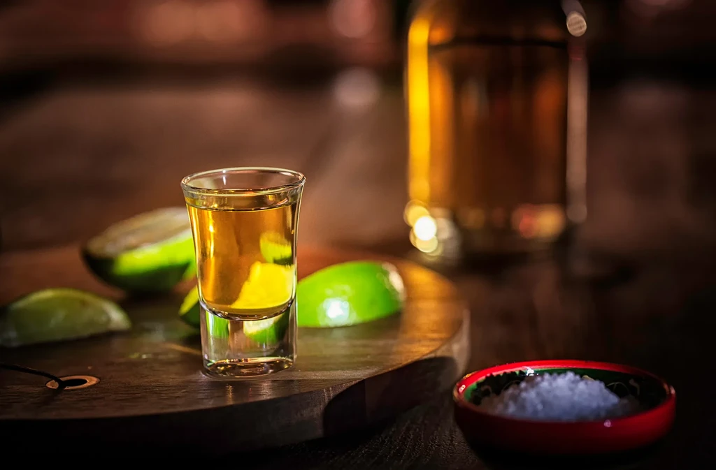 Glass of amber spirit, lime wedges, and salt on dark wood.
