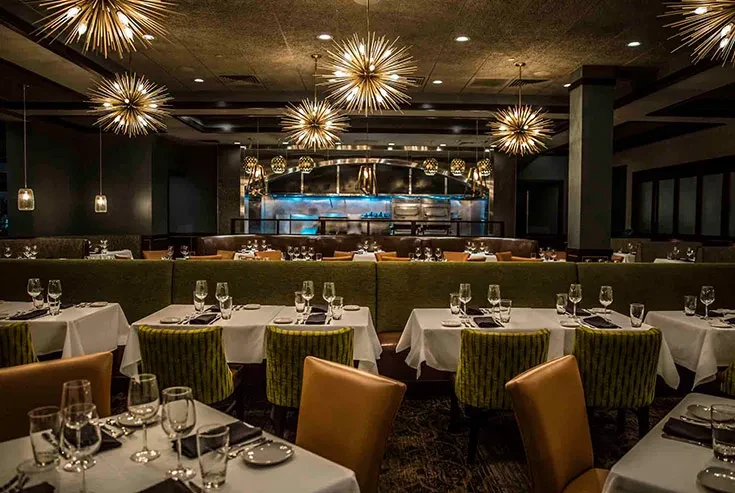 Elegant restaurant dining room with sputnik chandeliers, green booths, open kitchen.