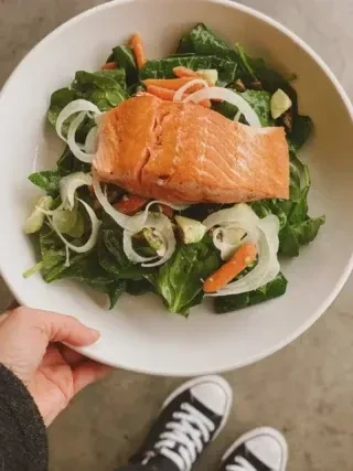 White bowl of salmon and green salad, person's hand and feet.