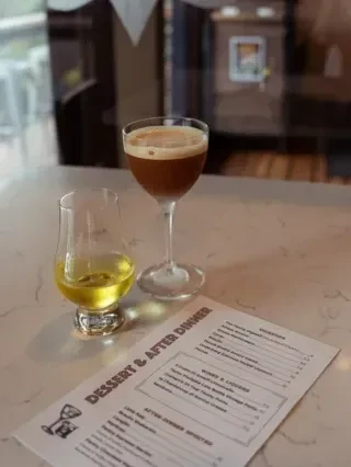 Nosing glass with golden spirit and dark foamy cocktail next to a dessert menu.