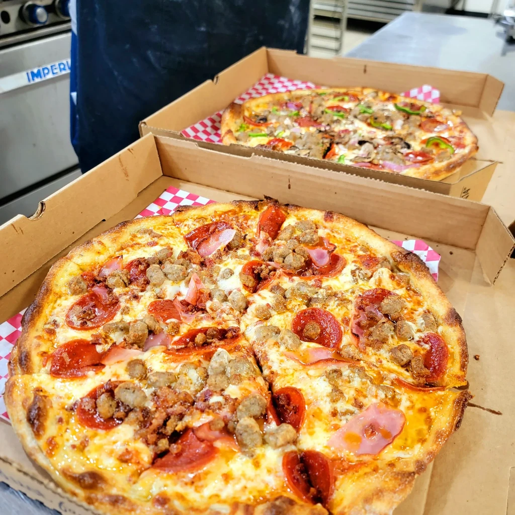 Two freshly baked pizzas, one meat, one veggie, in take-out boxes.