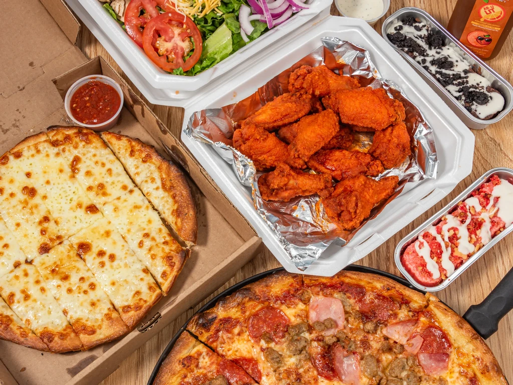 Flat lay of various takeout food items: pizzas, wings, salad, desserts, drink.
