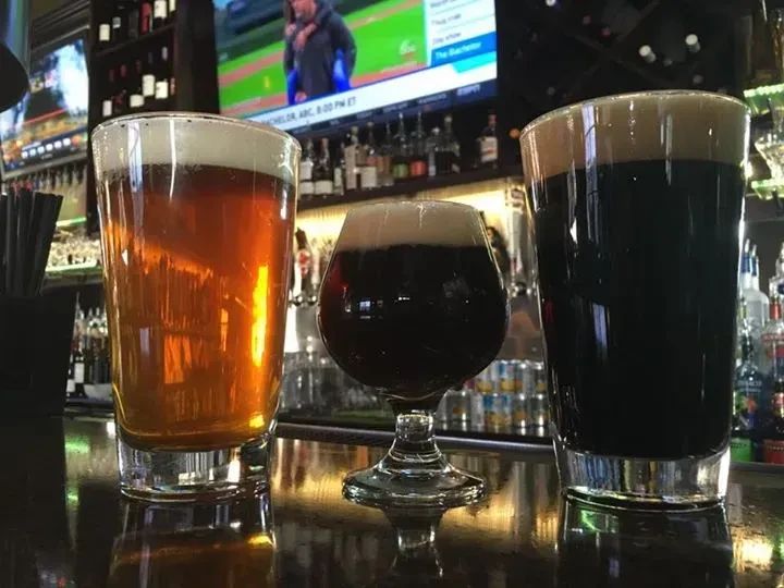 Three distinct beers on a reflective bar counter; TV in background.