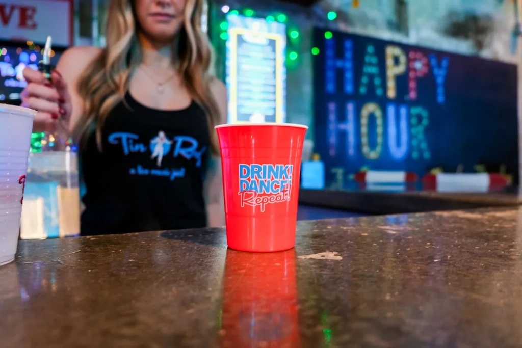 Red cup on bar, bartender wearing "Tin Roof" shirt mixing drink.