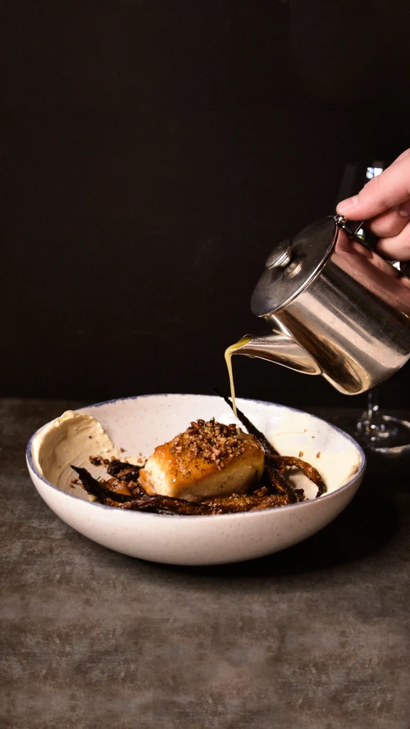 Hand pours yellow sauce onto golden food in a ceramic bowl.