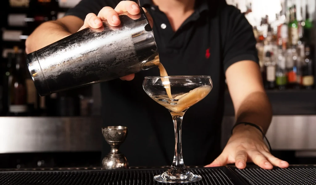 Bartender's hands pouring frothy cocktail from shaker into coupe glass.