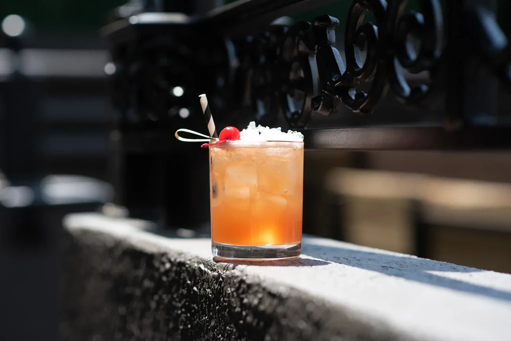 Amber cocktail with cherry, striped straw, and ice on an outdoor ledge.