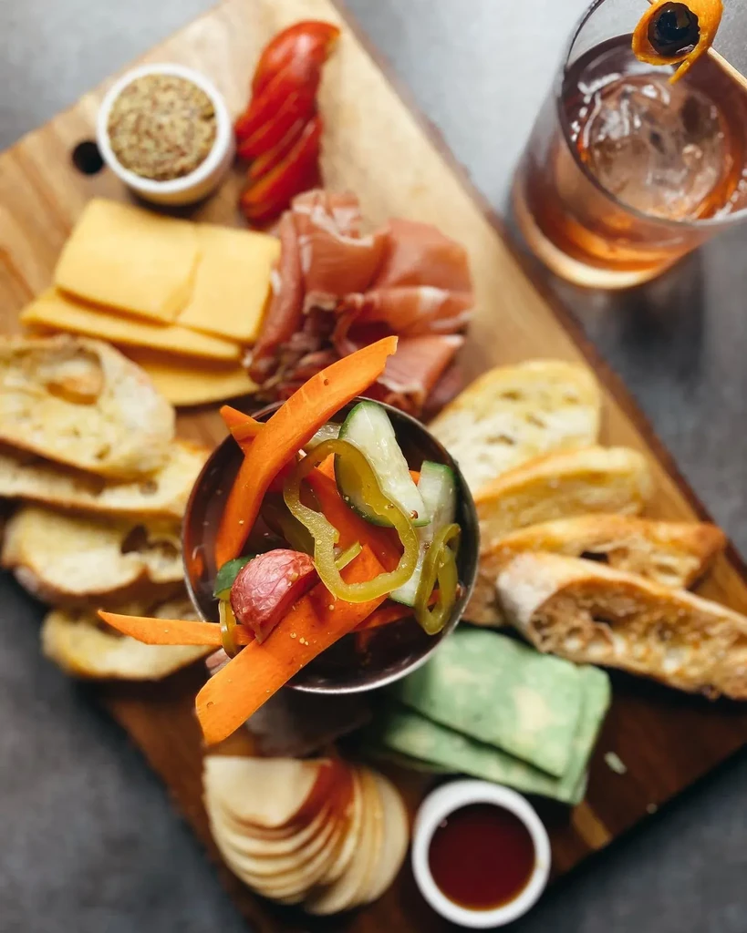 Charcuterie board with cheeses, meats, fruits, pickled vegetables, and a cocktail.