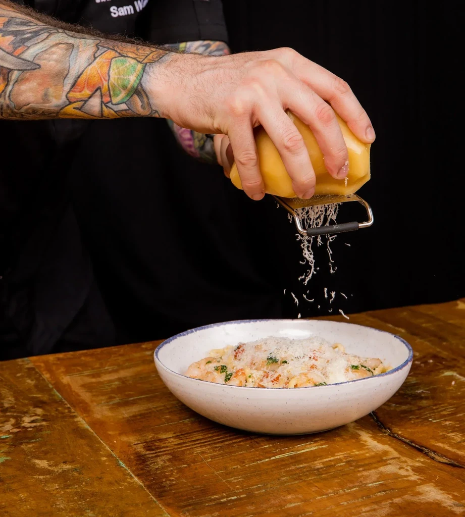 Close-up of tattooed hands grating cheese over a bowl of pasta.