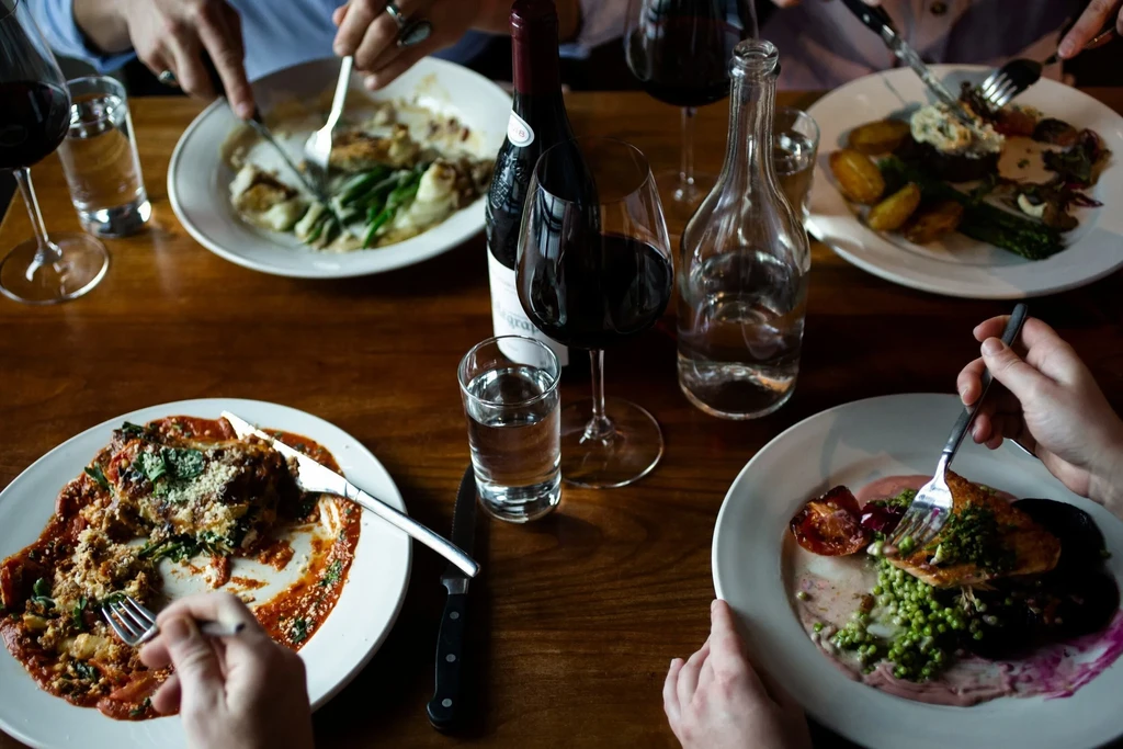 Multiple hands eating various meals and wine at a dark restaurant table.