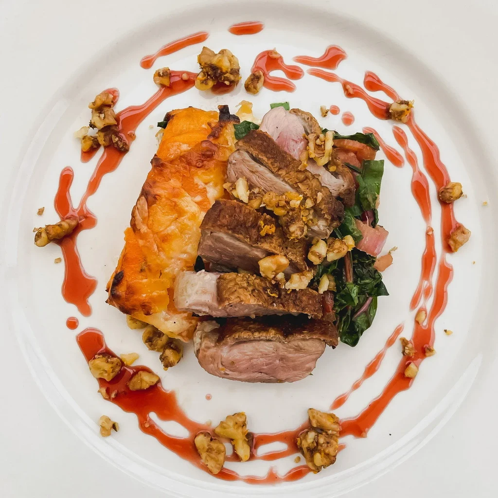 Plated meat, sweet potato, greens, walnuts, and red sauce.